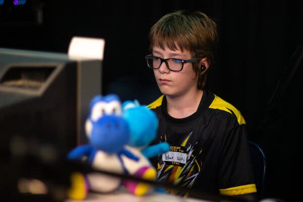 A 13-year-old boy with sandy-brown hair and black-framed glasses stares at a television screen as he plays a video game. He is wearing a name tag that says “blue scruti” in marker.