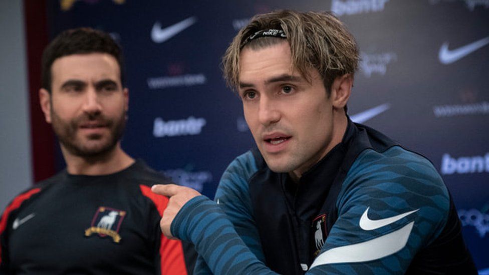 (L-R) Brett Goldstein as his character Roy Kent in a press conference with Phil Dunster as Jamie Tartt. Both are wearing football training tops and Jamie is speaking and pointing to Roy.