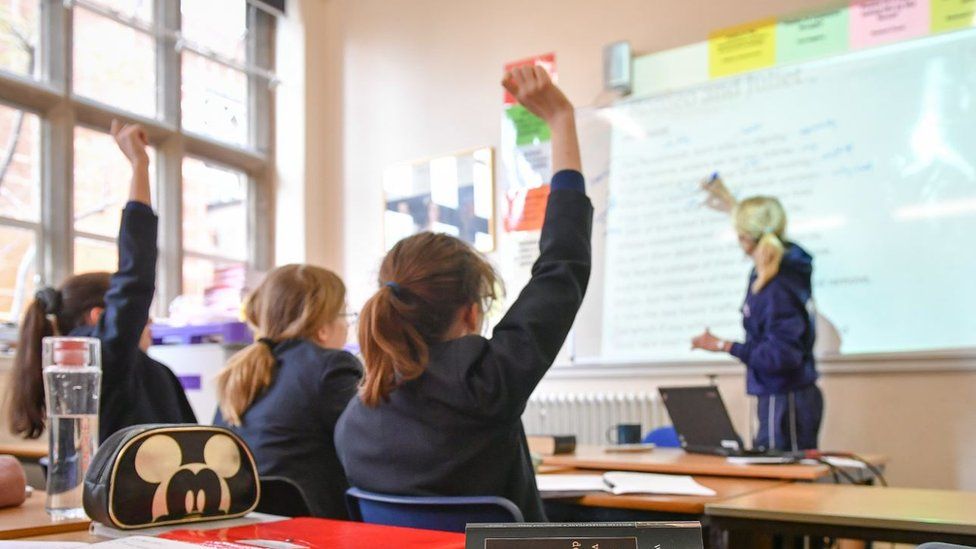 Students raise their hands as a teacher writes on a whiteboard in a classroom