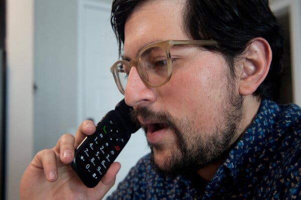 Jose Briones, wearing a blue patterned shirt and glasses, talking into a black flip phone.