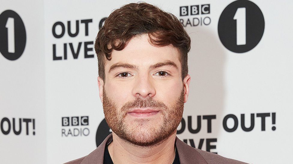 Jordan North pictured at a BBC Radio 1 event. Jordan is a white man in his 30s with short curly brown hair and a short brown heard. He has brown eyes and looks at the camera, wearing a brown suit jacket over a black top. Behind him is white and black Radio 1 branded event hoarding