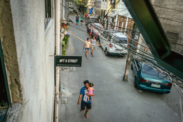 A man carrying a child walks past a building that has a small sign reading, “NFT X STREET.”