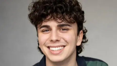 Melanie Gail Photography Sonny Monaghan. Sonny's a young man with dark curly hair and brown eyes. He smiles at the camera, wearing a blue and green striped polo shirt. 