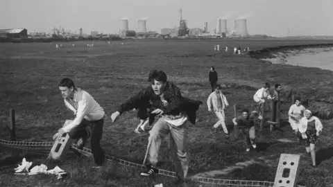 Getty Images Gatecrashers jump over the remains of the fence, with wateland and an industrial plant in the background