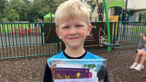 BBC Theo, a boy with blonde hair and a blue shirt holds up his books