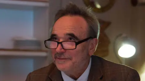 Chicken House Barry Cunningham wearing black glasses and a brown suit, looking off to the side