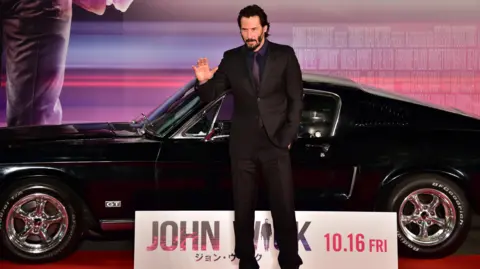 Getty Images Keanu Reeves, wearing a black suit, at a John Wick premiere in front of a black sports car