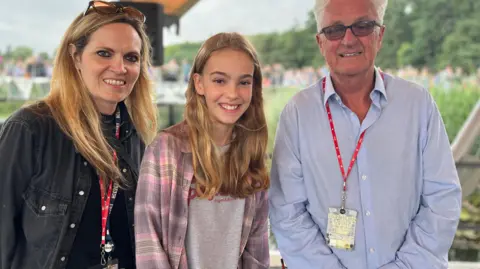 Jon Wright/BBC BBC Radio Suffolk's Sarah Lilley with long blonde hair and khol-rimmed eyes, Anna, with long blonde hair wearing a grey t-shirt and a pick plaid shirt and festival director Melvin Benn in a light blue shirt and short white hair and glasses standing on a stage