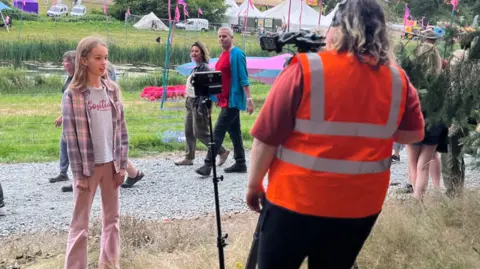 Jon Wright/BBC Anna stands in front of a TV camera with Latitude pink sheep in the background