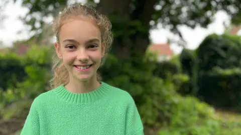 Jon Wright/BBC Anna, an 11 year old girl, wears a green jumper and stands in front of woodland