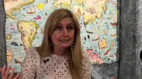 Cressida Cowell giving an interview in front of one of the exhibition's maps. Her hair is down and she wears a white flower top.