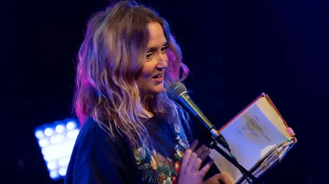 Wolverhampton Literature Festival Poet Hollie McNish on stage, she is talking into a microphone while holding a book, she is wearing a blue top with a pattern/graphic on the front