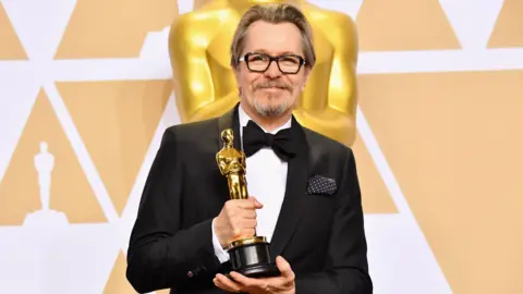 Getty Images Gary Oldman is wearing a black and white tuxedo with a black bow tie. He is holding his Academy Award trophy which is a golden statue of a man. 