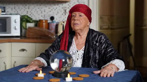  BBC / Kieron McCarron Laila Morse is wearing a red headband. She is sat at a table with a crystal ball and her hands are placed wither side of it. The crystal ball is surrounded by a circle of tea lights and biscuits.