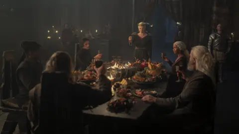 HBO Group of people in House of the Dragon, sat around an elaborate feast raising their glasses to toast