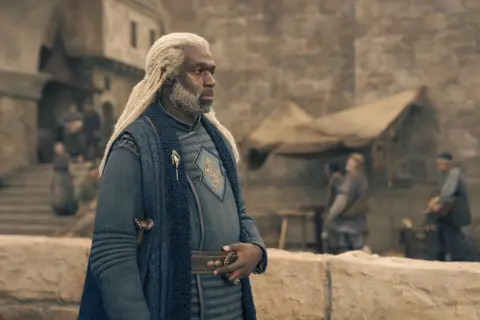 HBO A man with blonde dreadlocks wearing a blue period style costume 