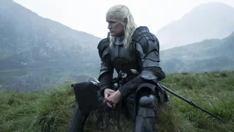 HBO A man with silver hair, sat on grass with mountains in background, wearing a suit of armor