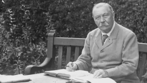 Getty Images A black and white image of Sir Arthur Conan Doyle sitting outdoors on a bench with an open set of notebooks and a pen in his hand