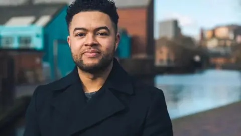 Paul Stringer A man in a black tailored coat standing in front of a stretch of the canal in Birmingham city centre