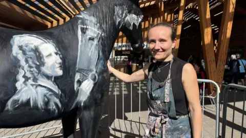 Andrew Turner/BBC Lois Cordelia standing next to the partially-completed painting on a model of a horse