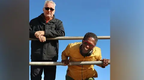 Richard Davis Henry Normal standing in front of railings and Lemn Sissay bending down and holding railings with blue sky