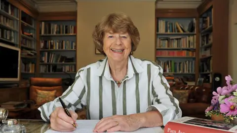 Mikal Ludlow Pam Ayres smiling and sitting at a desk in an office in a home writing as she writes in a book with a fountain pen in her hand and one of her books on the right of her desk