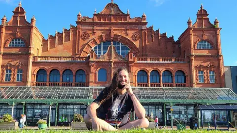 Matt Panesh/Morecambe Poetry Festival Matt Panesh sitting cross-legged on grass outside the Winter Gardens in Morecambe