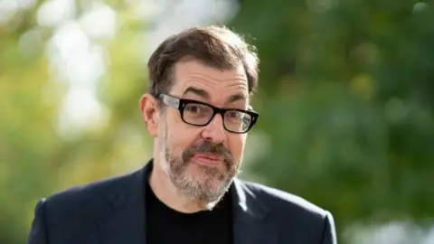 Getty A picture of Richard Osman. He is wearing a navy suit with a black t-shirt. The background is out of focus trees.