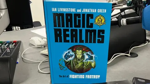 The Magic Realms The Art of Fighting Fantasy book stood up on a desk. It is a blue book with big, black letters. 