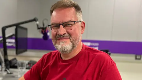 BBC Jonathan Green. He is pictured in a radio studio, sat at a desk with a mic. Jonathan has short light hair and facial hair. He is wearing glasses and a red t-shirt. He is looking at the camera and smiling.