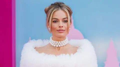 Getty Images A woman with blonde hair loosely tied back and artfully arranged poses for cameras. She's wearing a snug-fitting pearl necklace with three rows of beads, and a white, fluffy ruff-type accessory around her shoulders. 