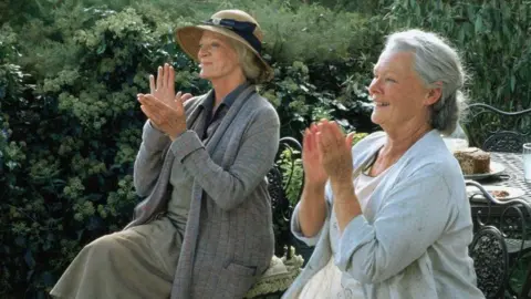 Alamy Dame Maggie Smith, left, and Dame Judi Dench, both wearing 1930s period dress, applaud and smile at something off camera - a table behind them has cake and drinks on it 