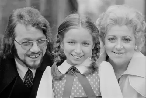 Getty Images Black and white photo of a young Lena Zavaroni standing in between her parents, Victor on the left and Hilda on the right. Her hair is in pigtails and she is wearing a spotted dress. Victor, who has glasses on, has a 70s-style haircut and a jacket and tie. Hilda is in a light jacket and pearl earrings and they are all looking at the camera.