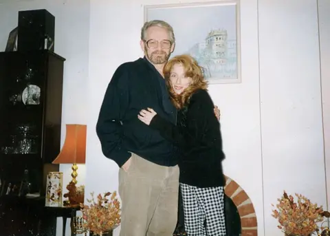 STAX/Specky Productions/BBC Scotland Victor Zavaroni, in beige trousers and a black cardigan, hugs his daughter who is wearing a black jumper and black and white checked trousers. She has long reddish brown hair and her face is drawn. They are in a sitting room beside a fireplace and table lamp.