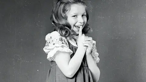 Getty Images A black and white photo of a young Lena Zavaroni smiling with her finger at her mouth. She is wearing a pinafore and a floral trimmed blouse with puff sleeves.