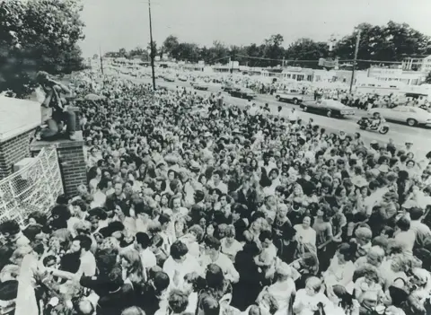 Getty Images A black and white image of thousands of people outside Graceland after Elvis died. You can see the fence of his property, and there is a photographer perched high on one of the bollards, taking photos of the huge crowds of people.