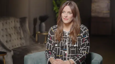 Riley Keough sits, slightly smiling for an interview in a living room. She has long brown hair and is wearing a colourful jacket with a white pattern on it. 