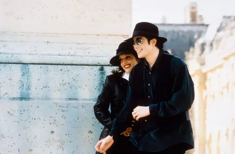 Getty Images Michael Jackson and Lisa Marie Presley in France walking past a stone building. They are both smiling, are both wearing dark clothes and both have black hats on. Michael has sunglasses on.