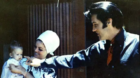 Getty Images Lisa Marie Presley as a baby being held by Priscilla Presley, who is looking at her smiling and wearing a large white bonnet-style hat. Elvis Presley is standing next to them, also smiling and pointing near Lisa's face, about to touch her nose. He's wearing a black shirt with red bandana around his neck.