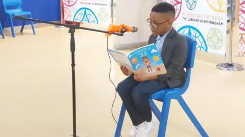 Anim Jayce sitting on a blue chair with his book A Beach With No Sea as he reads into a microphone that has been set up in front of him.