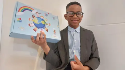 Anim Jayce, a boy with short dark hair and glasses, wearing a grey jacket and blue buttoned-up shirt while holding up a blue box with images of children on.