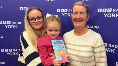 BBC Willow, pictured with her mum Rachel and author Sylvia Buck, holds a copy of a book entitled Willow Come to Tea? against a purple background.