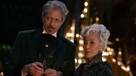 Universal Jeff Goldblum wears a dark suit and Michelle Yeoh is in an embellished top. They both have curious looks on their faces.