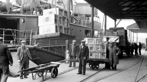 Bristol Archives A black and white photo of Bristol docks with a large container ship unloading large crates onto trolleys, with several men in flat caps in the foreground