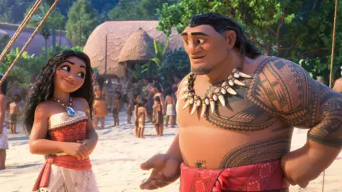 Disney A still from Moana 2, with the main characters on a Polynesian island, with lots of different characters on the beach in the background. There is a straw hut and lots of trees.