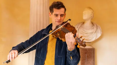 National Trust For Scotland Ewen Handerson performing with the Gregg fiddle. He's wearing a yellow jumper and blue jacket, while playing the fiddle. A white marble bust is behind him.