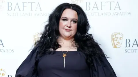 PA Ashley Storrie poses for the cameras at the Bafta Scotland Awards, wearing a black dress with puffed sleeves and an gold necklace.