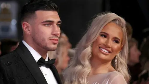 Getty Images Tommy Fury, wearing a black tuxedo with white shirt and black bow-tie, and Molly-Mae Hague, wearing a sheer dress and with long blonde hair, looking off camera - Molly-Mae is smiling 