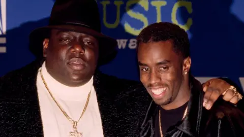 Getty Images Notorious BIG, wearing a black hat and gold chain with a crucifix, with his arm around a smiling P Diddy at the Billboard Music Awards in 1995