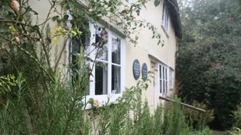 Essex Wildlife Trust Front of a yellow cottage with plants growing to the height of its window sills. Two round signs can be seen near the front door.